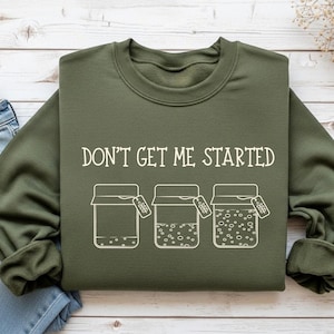 Może przedstawiać: Zielona bluza z napisem "DON'T GET ME STARTED" w kolorze białym. Poniżej tekstu znajdują się trzy słoiki z etykietami, każdy z różną zawartością. Bluza wykonana jest z miękkiego materiału.
