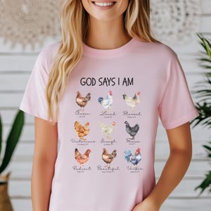 May include: Pink t-shirt with the text "God Says I Am" and a graphic of nine chickens with different colors and patterns. Each chicken has a word and a Bible verse underneath it. The words are: Chosen, Loved, Blessed, Victorious, Enough, Brave, Radiant, Beautiful, Unique.