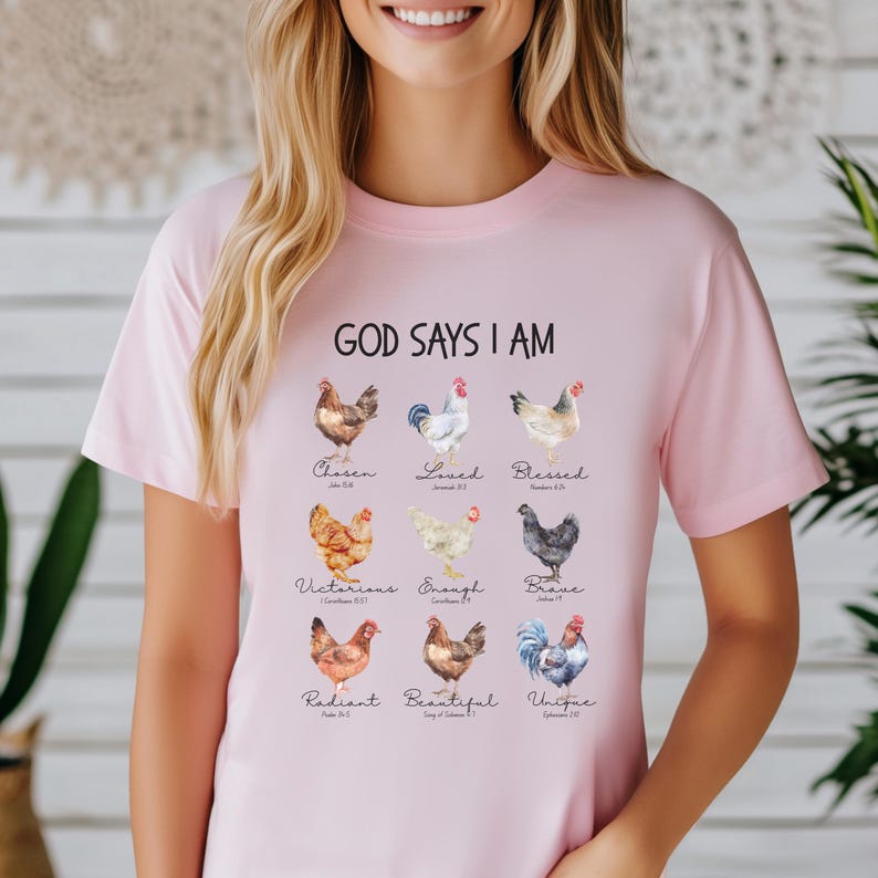 May include: Pink t-shirt with the text "God Says I Am" and a graphic of nine chickens with different colors and patterns. Each chicken has a word and a Bible verse underneath it. The words are: Chosen, Loved, Blessed, Victorious, Enough, Brave, Radiant, Beautiful, Unique.