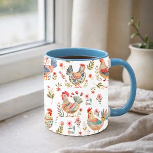 Puede incluir: Una taza de cer&aacute;mica blanca con interior y asa azules. La taza est&aacute; decorada con un patr&oacute;n repetido de gallinas coloridas, flores y follaje. La taza est&aacute; llena de un l&iacute;quido oscuro.