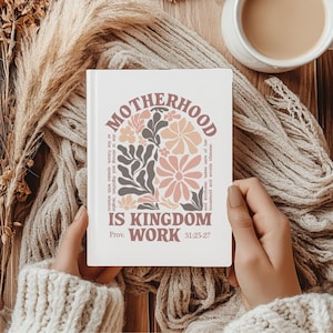 Könnte beinhalten: Ein weißes Notizbuch mit dem Text "MOTHERHOOD IS KINGDOM WORK" in einem floralen Design. Das Notizbuch wird von einer Person gehalten, die einen cremefarbenen Pullover trägt. Das Design umfasst Blumen in Rosa-, Pfirsich- und Brauntönen.