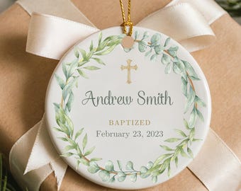 Może przedstawiać: Biała ceramiczna ozdoba ze złotym krzyżem i imieniem "Andrew Smith". Ozdoba jest ozdobiona zielono-białym wieńcem z liści. Słowo "BAPTIZED" i data "February 23, 2023" są wydrukowane. Złoty sznurek do zawieszenia.