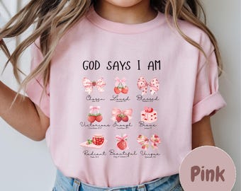 Puede incluir: Camiseta rosa con el texto "GOD SAYS I AM" y varias palabras e ilustraciones inspiradoras. El diseño incluye lazos, fresas y palabras como "Chosen", "Loved" y "Blessed".