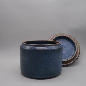 Pode incluir: Um pote de cerâmica azul escuro com uma tampa a condizer. O pote é cilíndrico com uma superfície ligeiramente texturizada. A tampa é redonda com uma borda castanha. O pote e a tampa estão sobre um fundo cinzento.
