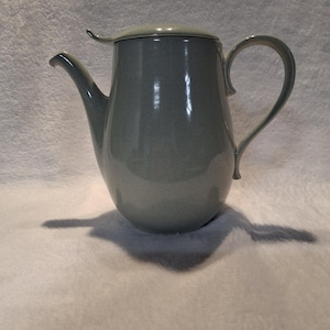 May include: A light gray ceramic teapot with a speckled finish, featuring a curved handle, a tapered spout, and a matching lid. The teapot has a smooth, glossy surface and a rounded body shape, presenting a clean and functional design for serving.