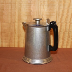 May include: A vintage metal coffee pot with a lid and a black handle. The pot has a silver finish and a rounded base. The background is a light brown wooden wall and a brown surface.