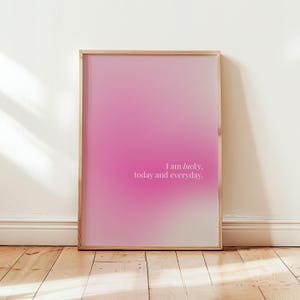 May include: A framed print with a pink and white gradient. The artwork displays the text "I am lucky, today and everyday" in a light font. The frame is light wood, and the print is against a white wall and wooden floor.