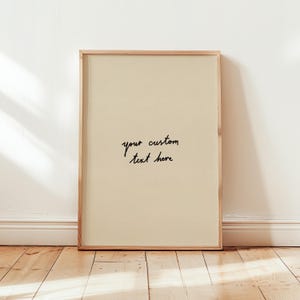 Op de afbeelding: Een ingelijste print met een lichtbeige achtergrond en de handgeschreven tekst "your custom text here" in zwart. De lijst is gemaakt van lichtgekleurd hout en de print leunt tegen een witte muur op een houten vloer.