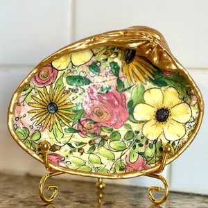 May include: A decorative seashell dish with a gold-painted rim and stand. The interior features a floral design with yellow, pink, and green flowers. The dish is displayed on a gold metal stand.