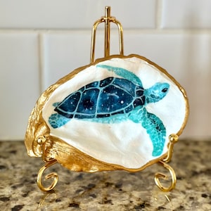 May include: A hand-painted sea turtle on a white oyster shell, displayed on a small gold easel. The turtle is depicted in shades of blue and teal, with a starry pattern on its shell. The shell has a gold-painted edge.