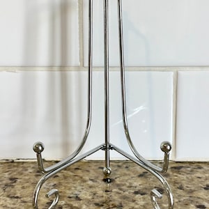 May include: A silver-colored metal plate stand with a three-legged base and a curved design. The stand has a vertical support with a small ball at the top and two small balls at the base of the curved legs. The stand is on a speckled brown surface.