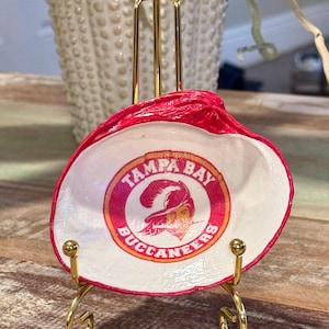 May include: A painted seashell with a Tampa Bay Buccaneers logo, displayed on a small gold stand. The shell is white inside with a red rim and the team logo in the center.