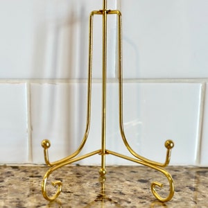 May include: A gold-colored metal display stand with a three-legged base and a vertical support. The stand has a decorative, curved design at the base and a small ball at the top. The stand is designed to hold a plate or other decorative item.