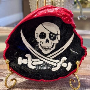 May include: A pirate-themed decorative piece featuring a skull and crossbones design. The artwork is painted on a black background with white accents and a red border. It is displayed on a gold-colored stand.