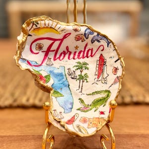 May include: A decorative oyster shell with gold-painted edges, displayed on a gold metal stand. The shell features a hand-painted map of Florida, the word Florida in pink script, a sun, palm trees, sailboats, a dolphin, and an alligator.