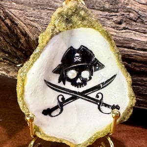 May include: A pirate-themed oyster shell decoration. The shell is painted with a black skull and crossbones design, wearing a pirate hat. The shell is mounted on a gold-colored stand. The shell has a gold-colored border.