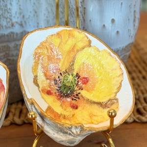 May include: A decorative oyster shell dish featuring a hand-painted yellow poppy flower with a green center and gold-painted edges, displayed on a small gold-toned metal stand. The interior of the shell is white with a glossy finish.