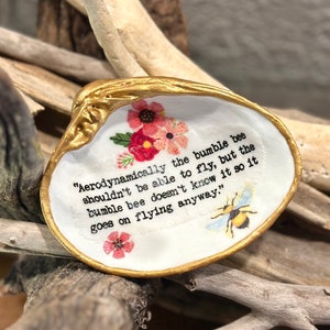 May include: A decorative seashell with a gold-painted rim and a white interior. The shell features a floral design and a quote about bumblebees. The quote reads: "Aerodynamically the bumble bee shouldn't be able to fly..."