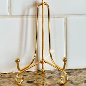 May include: A gold-colored metal display stand with a three-legged base and a vertical support. The stand has a decorative, curved design and is suitable for displaying plates or artwork. The stand is made of metal.
