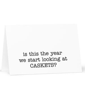 May include: A white greeting card with the text "is this the year we start looking at CASKETS?" in black. The card is folded and standing upright, with a white background.