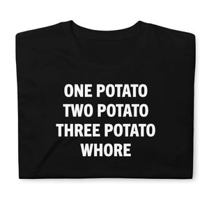 May include: A black short-sleeved t-shirt featuring white text printed on the front. The text is arranged in four lines and reads: ONE POTATO, TWO POTATO, THREE POTATO, WHORE. The shirt has a crew neckline and is displayed folded flat.