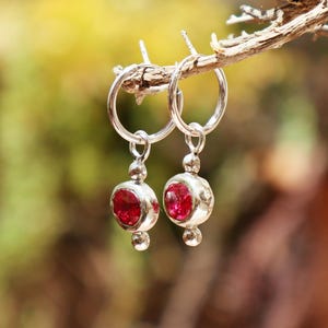 Puede incluir: Un par de pendientes de aro de plata con colgantes circulares que presentan piedras de color rojo intenso facetadas. Cada colgante está decorado con pequeñas esferas de plata en la parte superior e inferior, suspendidos de aros finos.