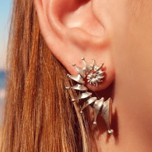 Peut inclure: Boucles d'oreilles sculpturales argentées avec un motif en spirale et un petit accent rouge. Les boucles d'oreilles ont une construction unique en couches avec une série d'éléments en forme de cône. Les boucles d'oreilles sont portées sur une oreille.
