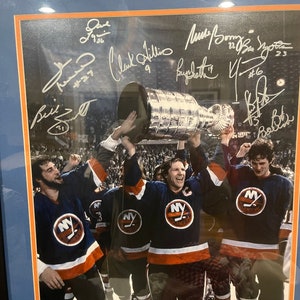 May include: A framed photograph of a group of hockey players holding up the Stanley Cup trophy. The players are wearing blue and orange jerseys with the New York Islanders logo. The photo is signed by several players, including Mike Bossy, Clark Gillies, and Butch Goring.