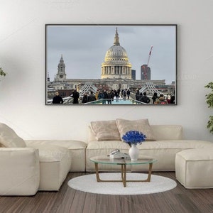 Puede incluir: Una impresión enmarcada de la Catedral de San Pablo en Londres, con su icónica cúpula y arquitectura circundante. La imagen captura un día nublado con gente caminando por un puente. La impresión se exhibe sobre un sofá seccional color crema.