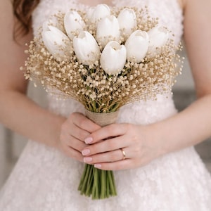 Peut inclure: Un bouquet de mariée composé de tulipes blanches et de délicats gypsophiles. Les fleurs sont maintenues ensemble avec de la ficelle et des tiges vertes. Le bouquet est tenu par une personne portant une robe en dentelle blanche.
