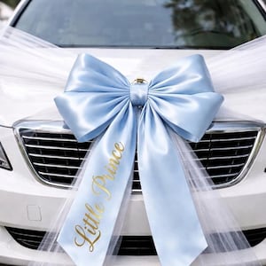 May include: A large, light blue satin bow attached to the front of a white car. The bow features long, flowing white tulle fabric extending to the sides. The left tail of the blue bow has the words Little Prince printed in elegant gold script.