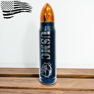 May include: A navy blue bullet-shaped tumbler with a gold-colored top. The tumbler has the letters "USMC" engraved on the side, along with a bulldog graphic. The American flag and the text "THE PATRIOT GUY" are in the background.