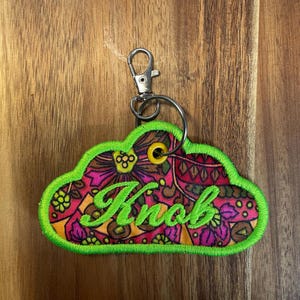 May include: Embroidered cloud-shaped keyring with a vibrant floral pattern in pink, orange, and brown. The word "Knob" is embroidered in green script. The keyring has a bright green border and a silver clasp.