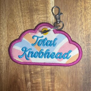 May include: A cloud-shaped keyring with the words "Total Knobhead" embroidered in blue. The keyring has a pink border and a colourful striped background in shades of pink, orange, and blue. It includes a silver-coloured clasp.
