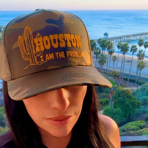 Może przedstawiać: Czapka typu trucker w kolorze moro z napisem "HOUSTON I AM THE PROBLEM" w złotych literach i grafiką kaktusa. Czapka ma siatkowy tył i zakrzywiony daszek.