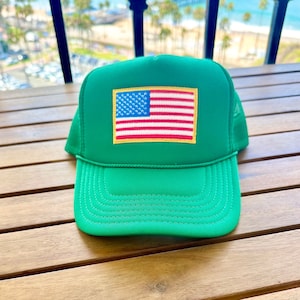 Puede incluir: Una gorra de camionero verde con una parte trasera de malla y un pequeño parche de la bandera estadounidense en la parte delantera.