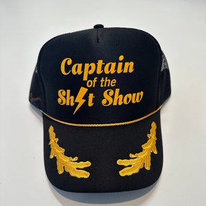 May include: Black trucker hat with a gold embroidered lightning bolt and the text "Captain of the Sh*t Show".