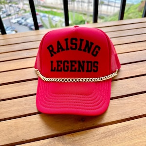 May include: Red trucker hat with a black mesh back and a gold chain. The hat has the text "Raising Legends" in black.