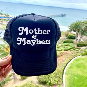 May include: Black trucker hat with a white mesh back. The hat has white text that reads "Mother of Mayhem" in a retro font.