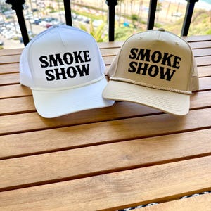 May include: Two trucker hats with the text "SMOKE SHOW" in black lettering on a white and tan hat.