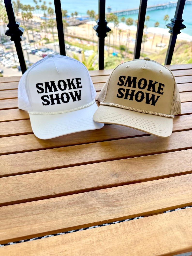 May include: Two trucker hats with the text "SMOKE SHOW" in black lettering on a white and tan hat.