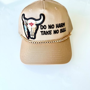 Puede incluir: Gorra de camionero color canela con detalle de cadena dorada y un parche con una calavera de toro con plumas y el texto "DO NO HARM TAKE NO BULL". La gorra tiene una parte trasera de malla.