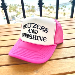 May include: A pink and white trucker hat with the text "Seltzers and Sunshine" in black embroidery.