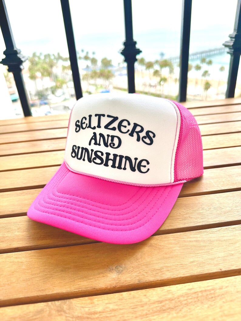 May include: A pink and white trucker hat with the text "Seltzers and Sunshine" in black embroidery.