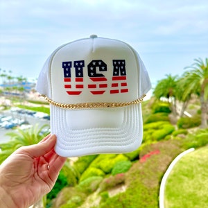 Pode incluir: Boné de caminhoneiro branco com um gráfico dos EUA vermelho, branco e azul e uma corrente dourada ao redor da borda.