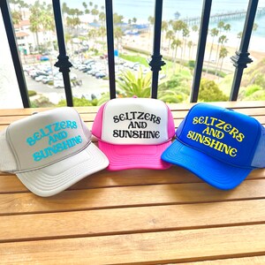 May include: Three trucker hats with mesh backs. The hats are gray, pink, and blue. Each hat has the text "Seltzers and Sunshine" embroidered on the front in a white and turquoise font.