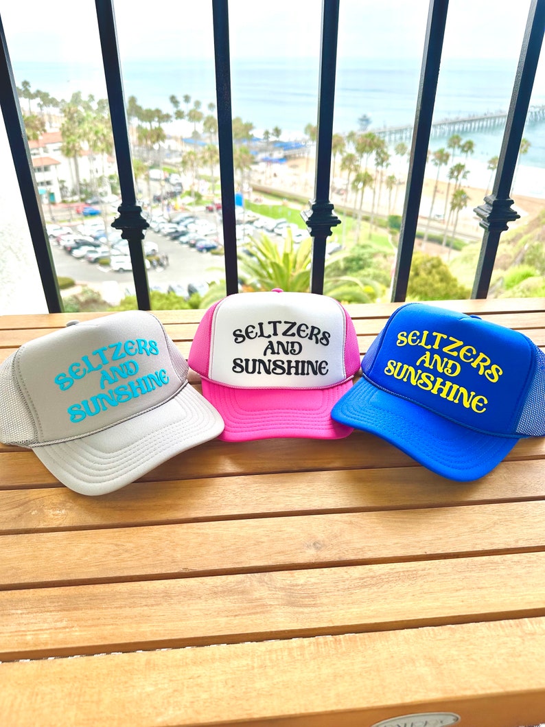 May include: Three trucker hats with mesh backs. The hats are gray, pink, and blue. Each hat has the text "Seltzers and Sunshine" embroidered on the front in a white and turquoise font.