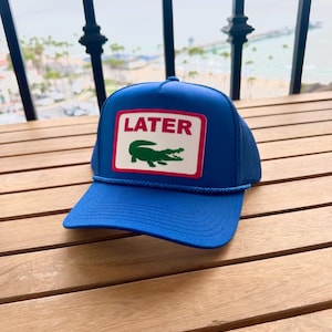 May include: A blue trucker hat with a white and red rectangular patch. The patch features the word "LATER" above a green crocodile logo. The hat has a blue brim and a mesh back.