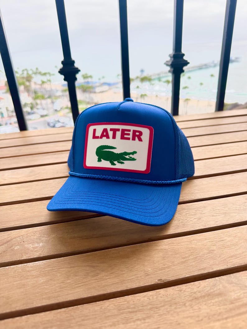 May include: A blue trucker hat with a white and red rectangular patch. The patch features the word "LATER" above a green crocodile logo. The hat has a blue brim and a mesh back.