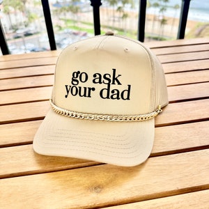Könnte beinhalten: Ein beiger Trucker-Hut mit einer goldenen Kette um den Rand. Der Hut hat einen schwarzen gestickten Text, der "go ask your dad" lautet.
